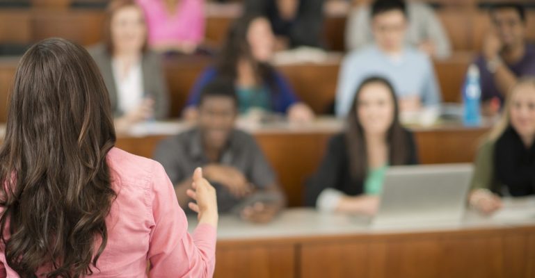 A multi-ethnic group of college age students are sitting in a row in a lecture hall and are listening to their professor.