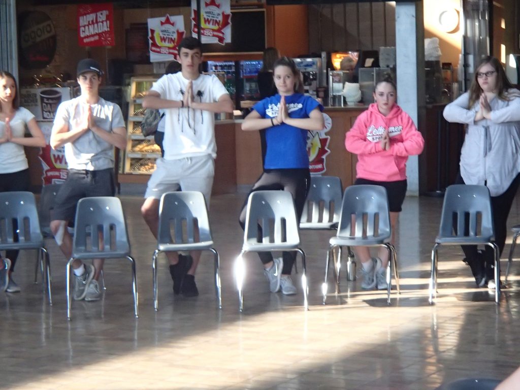 Campers balance on one leg in yoga pose behind chairs in Agora