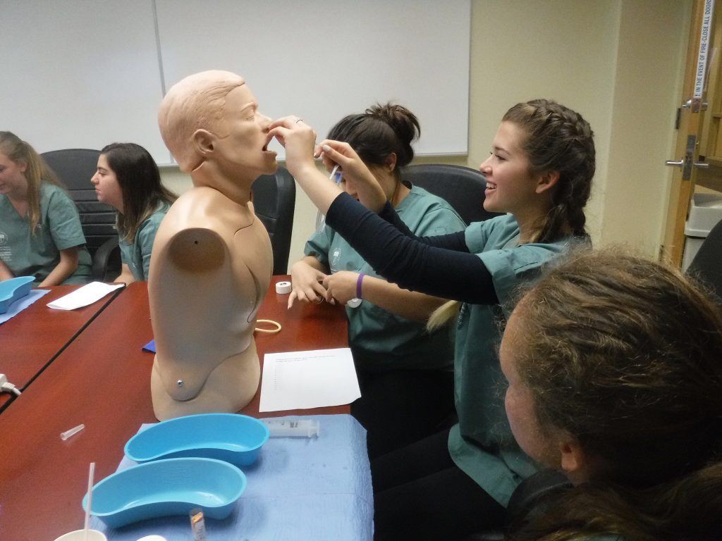 Campers sit around table in small group room while one camper practices inserting a naso-gastric tube into a SimMan mannequin bust