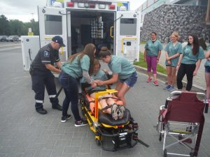 Campers practice strapping fellow camper onto EMS stretcher outside of medical school building while paramedic assists