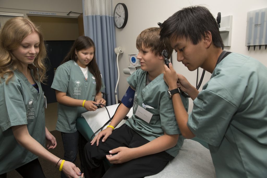 Campers practicing multiple clinical skills; one taking blood pressure, one examining inner ear, and one testing knee reflexes
