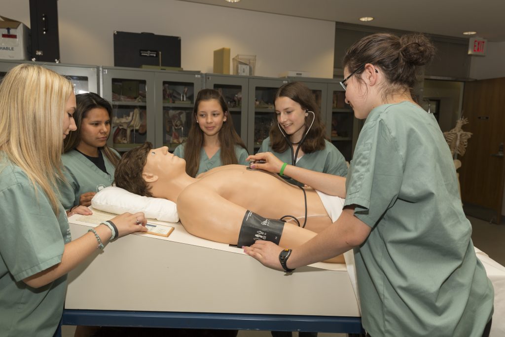 Camper practicing using a stethescope with SimMan 3G mannequin while fellow campers look on
