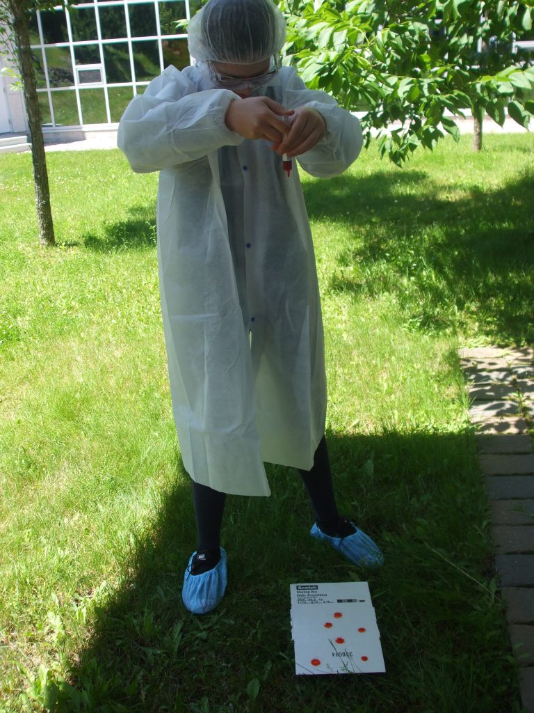 Camper wearing CSI coat, cap, glasses, and booties uses syringe to drop fake blood drops onto blood spatter analysis card
