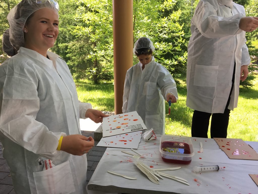 Camper laughs at camera while holding blood spatter analysis cards. Two fellow campers in background uses syringe to create blood drops from different heights on a blood spatter analysis card outside