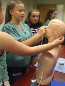 Camper places naso-grastric tube into SimMan mannequin bust while two fellow campers look on in small group room