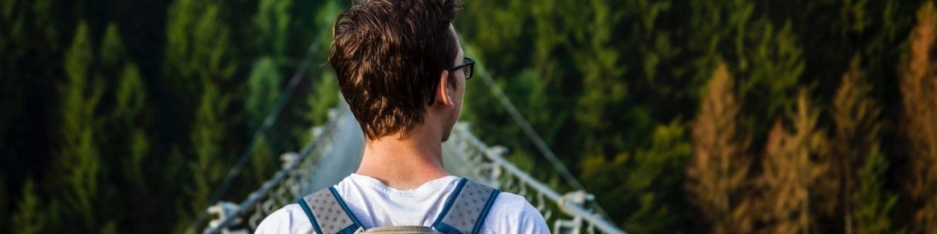 Research Homepage. Young Man wearing backpack walking away from viewer across a suspension bridge. Evergreen Trees in the background.