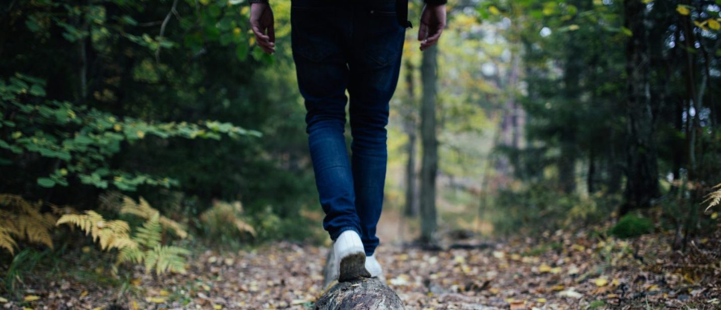 Person wearing jeans walking along a trail in the forest surrounded by trees. Can only see the individual from the legs down.