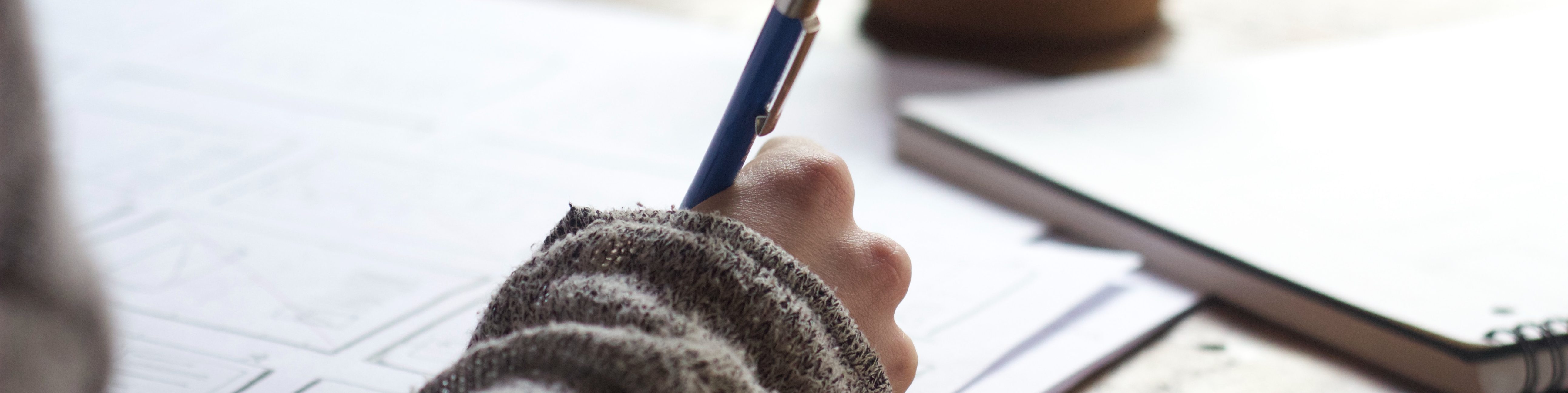 An individual editing a paper on a desk holding a pen. The corner of a notebook is visible in the top right corner