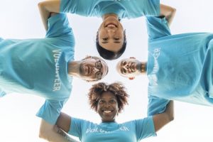 Four individuals wearing blue volunteer t-shirts linking arms in square shape and looking down at camera