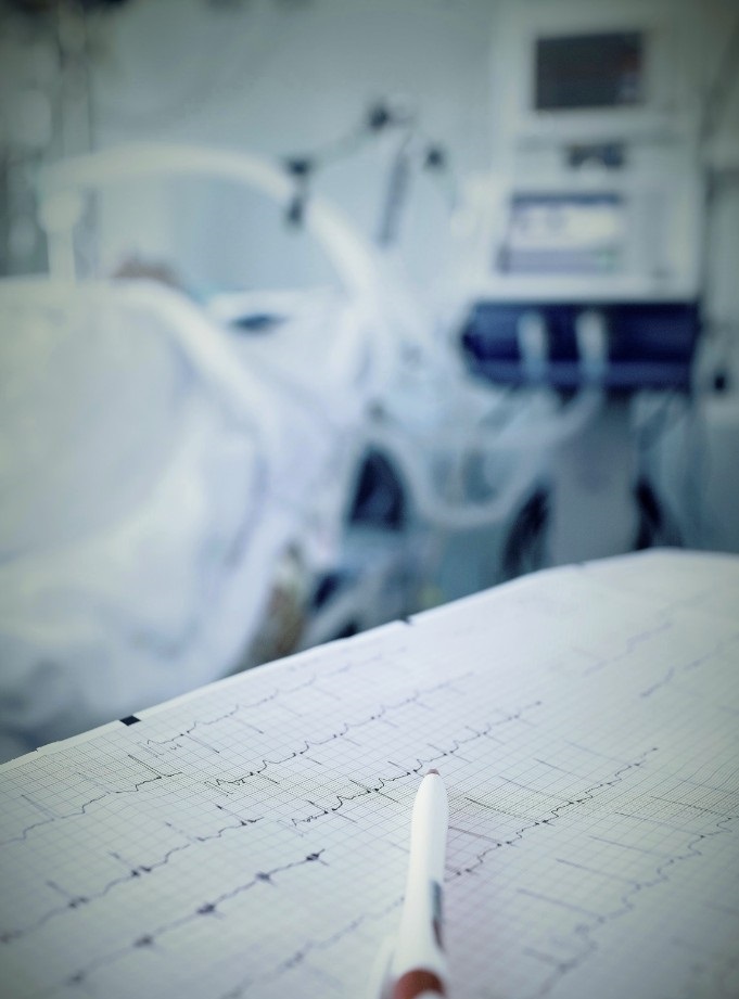 Photo of cardiograph results with a pen resting on the paper. Blurred image of a patient on a stretcher in the background.