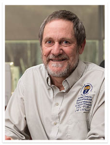 Photo of Dr. Roger sitting in his office, at a desk. Dr. Strasser is wearing a light blue shirt with a NOSM logo embroidered on the left chest.
