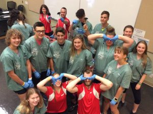 Campers and team leads strike funny pose while wearing blue gloves in classroom; 2 team leads and 1 camper use hands to make owl eyes over their eyes
