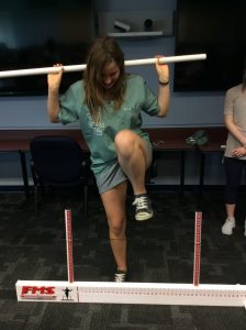 Camper steps over string tied between two measuring poles approximately one foot from the ground while holding a balance bar across their shoulders