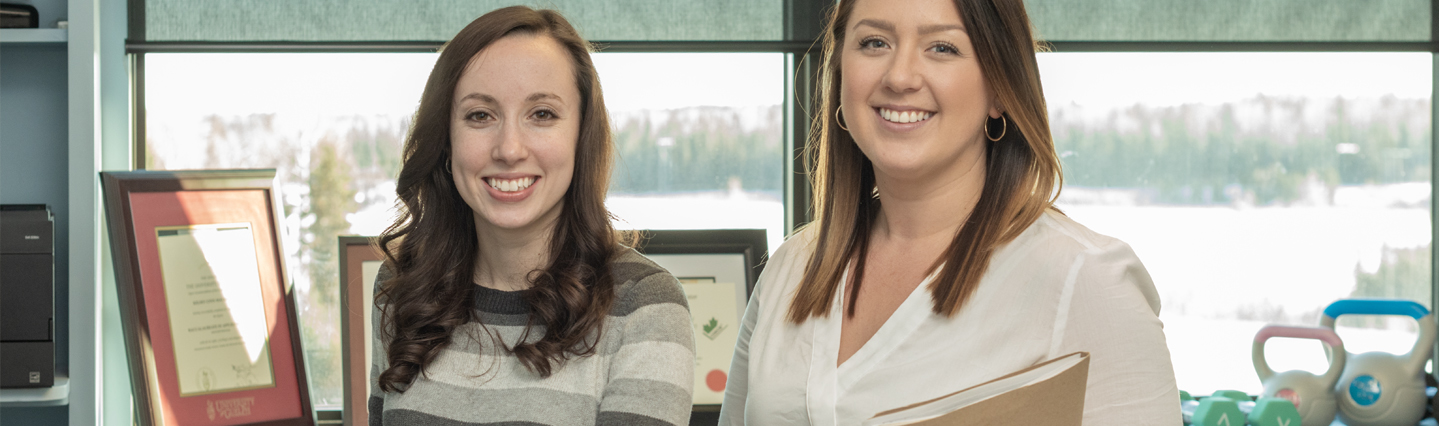 Two women wearing white and grey stand in an office in front of a window in winter