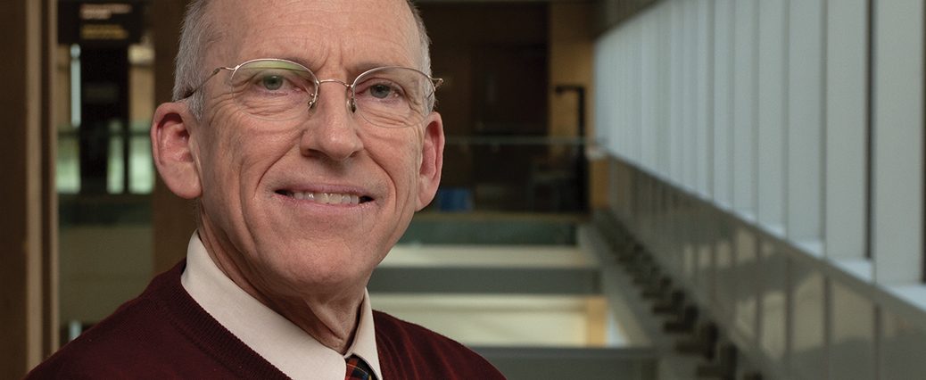 older man with glasses wearing red sweater stands smiling in front of empty hallway