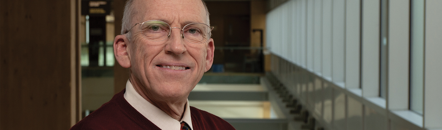 older man with glasses wearing red sweater stands smiling in front of empty hallway