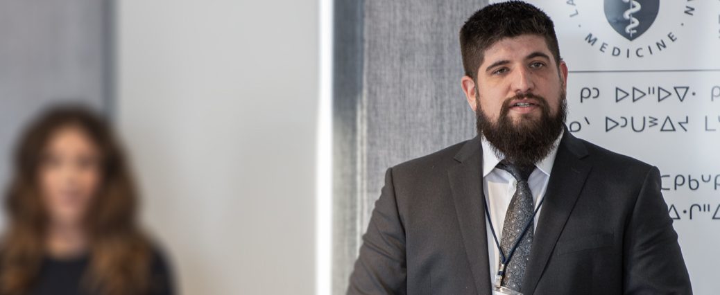 Man with dark hair and beard gives presentation in front of NOSM banner