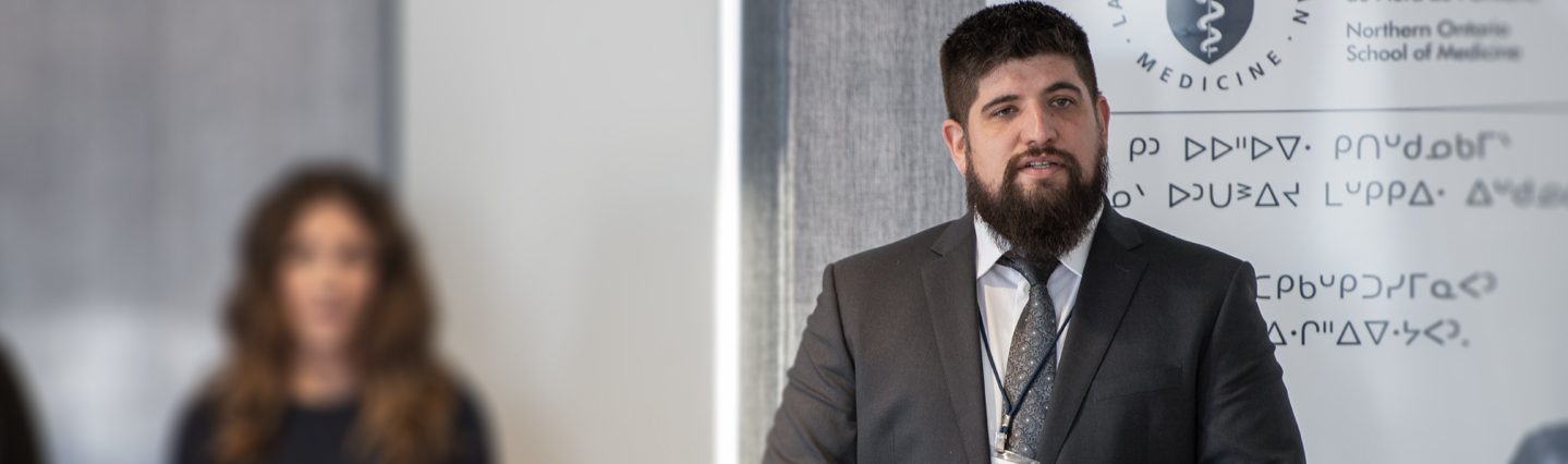 Man with dark hair and beard gives presentation in front of NOSM banner