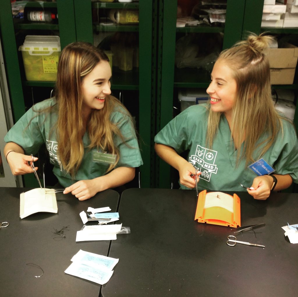 Two campers laugh while practicing suture skills on suture kits in lab classroom.