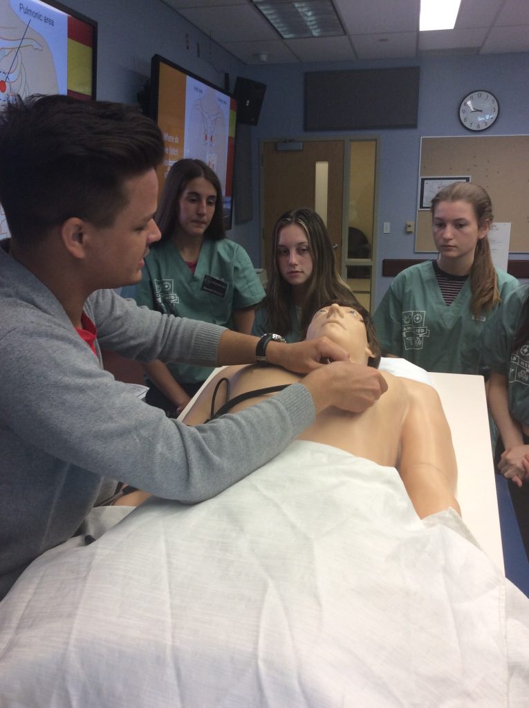 Team lead demonstrating how to listen to heart sounds with stethoscope on simulation mannequin.
