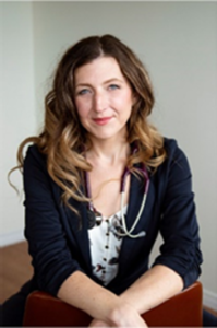 Dr. Melanie Squarzolo is smiling while sitting on a chair backwards, wearing a blue blazer jacket and white flowered shirt 