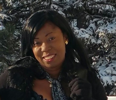 Dr. Blessing Odia is smiling outside in front of snow covered pine trees, wearing a black shirt and black scarf 