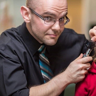 Dr. Lyall Pacey is looking at camera while checking patient's ear, wearing glasses, a black shirt and striped tie 