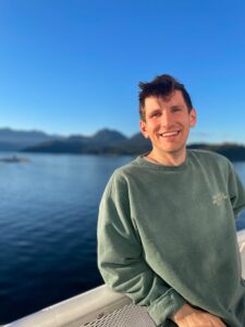 Dr. Cole Kubay is smiling wearing a green sweatshirt, standing in front of a body of water 