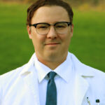 Dr. Matthew Prisk is smiling wearing glasses and a doctor coat with white collard shirt and blue tie