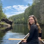 Dr. Nicole Brunet is smiling wearing a black shirt, looking backward while sitting on a canoe in a river