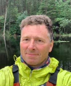 Dr. Patrick Martel is smiling wearing a yellow rain jacket in front of a body of water