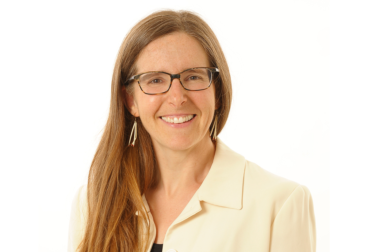 Dr. Erin Cameron wears a cream jacket, glasses, and long gold earrings. She is smiling and has long, straight strawberry-blonde hair. Erin Cameron, Ph.D., porte une veste crème, des lunettes et de longues boucles d’oreilles en or. Elle est souriante et a de longs cheveux raides d’un blond roux.
