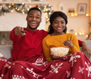 Favorite movie together at home and self-isolation during lockdown. Smiling young african american lady with popcorn, guy with remote control, covered with blanket watch film in interior with garlands