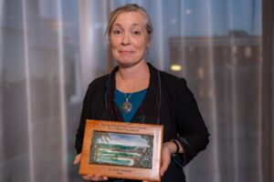 Dr. Sarah Newbery holds up a framed plaque. She wears a black jacket over a teal shirt and a large silver necklace. 