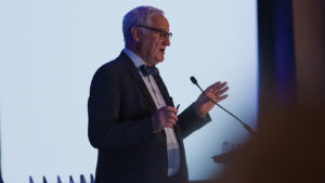Dr. Bill McCready stands at a microphone and gestures with his hands. A screen is behind him. He wears a black suit jacket, a bow tie, and glasses.