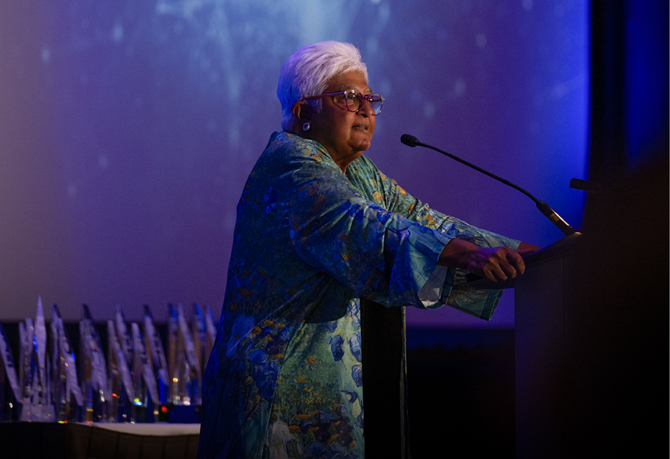 Dr. Sarita Verma stands in front of a podium and talks into a microphone. Behind her is a table with dozens of awards and a screen displaying stars.