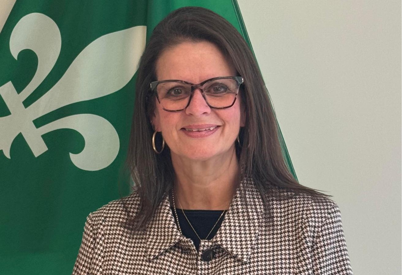 Danielle Barbeau-Rodrigue stands next to the Franco-Ontarian flag. She has long brown hair and wears glasses and a houndstooth jacket.