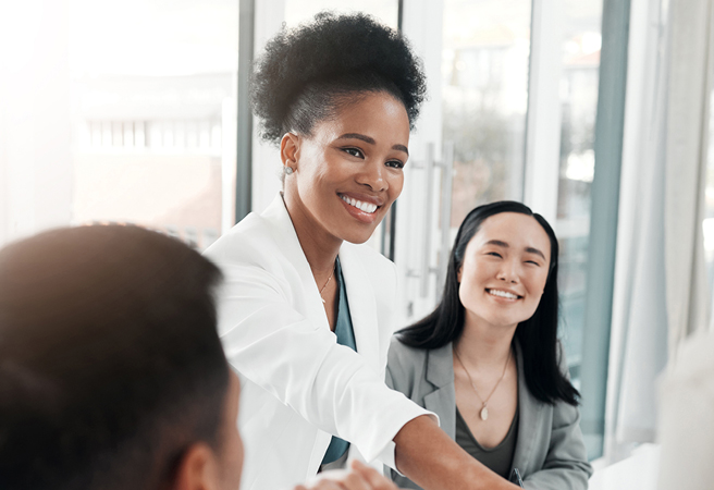 A young professional woman reaches forward in a handshake with another than cannot be seen. There are others around her.