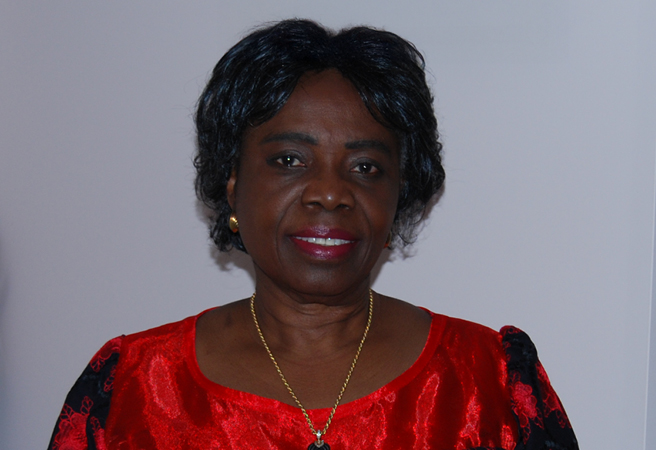 Dr. Remi Ogundimu sits in her living room and wears a red and black floral dress. La Dre Remi Ogundimu est assise dans son salon et porte une robe fleurie rouge et noire.