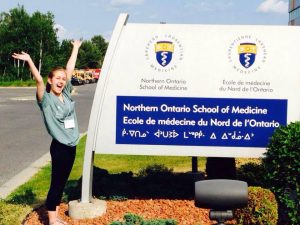Alyssa Labelle se tient près d’un ancien panneau indicateur de l'« École de médecine du Nord de l'Ontario ». Elle a un grand sourire et lève les bras en signe de joie.