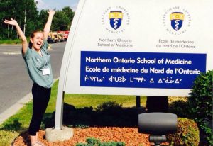 Alyssa Labelle se tient près d’un ancien panneau indicateur de l'« École de médecine du Nord de l'Ontario ». Elle a un grand sourire et lève les bras en signe de joie.