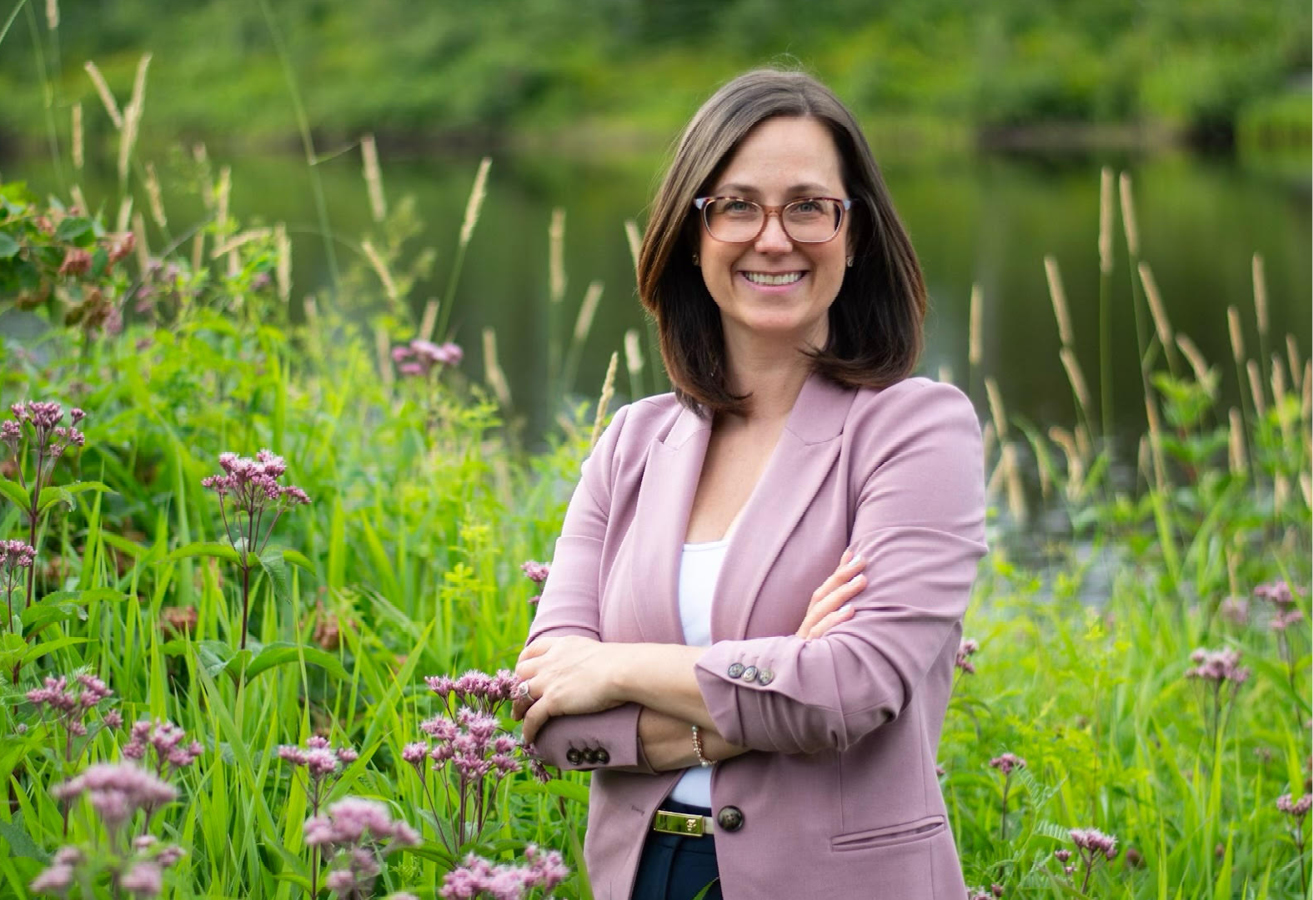 Dr. Marjolaine Talbot-Lemaire stands in a field of flowers with a river behind her. La Dre Marjolaine Talbot-Lemaire se tient dans un champ de fleurs traversé par une rivière.