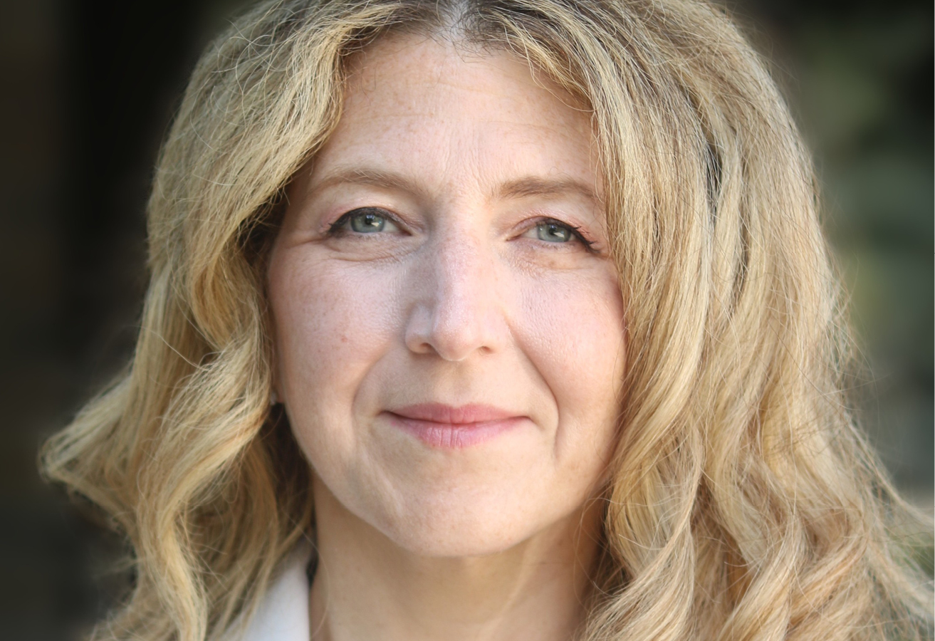 Dr. Melanie Squarzolo wears a white blazer. She has long curly blonde hair. La Dre Melanie Squarzolo porte une veste blanche. Elle a de longs cheveux blonds bouclés.