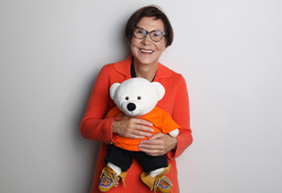 Dr. Cindy Blackstock stands holding Spirit Bear in front of her. Spirit Bear is a white stuffie wearing an orange t-shirt, black pants, and moccasins. Dr. Blackstock wears an orange jacket and glasses. Mme Cindy Blackstock, Ph.D., est debout et tient l’ours esprit. L’ours esprit est un ours blanc en peluche qui porte un t-shirt orange, un pantalon noir et des mocassins. Mme Blackstock porte une veste orange et des lunettes.